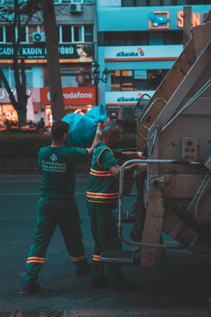 Two waste collection workers are engaged in rubbish removal on a city street during the evening, with illuminated commercial signage in the background. The worker on the left is standing, reaching upw