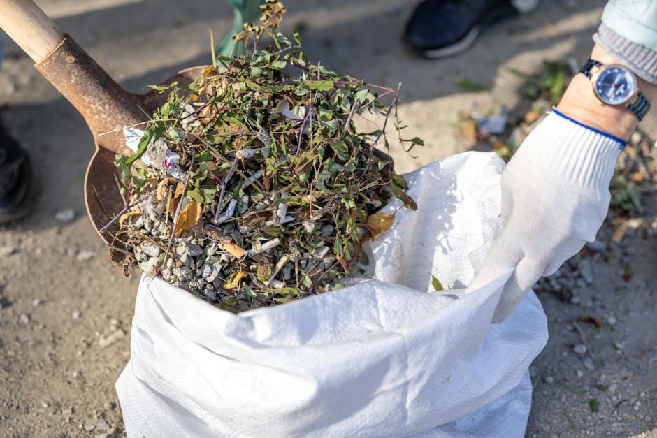 A close-up view of a white woven polypropylene sack filled with organic waste, primarily consisting of leafy branches, small twigs, and plant debris, partly covered with soil and small stones. The sac